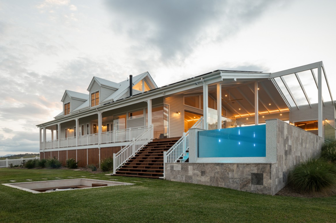 Queensland weatherboard home at dusk with elevated pool featuring glowing turquoise acrylic window panel in stone retaining wall